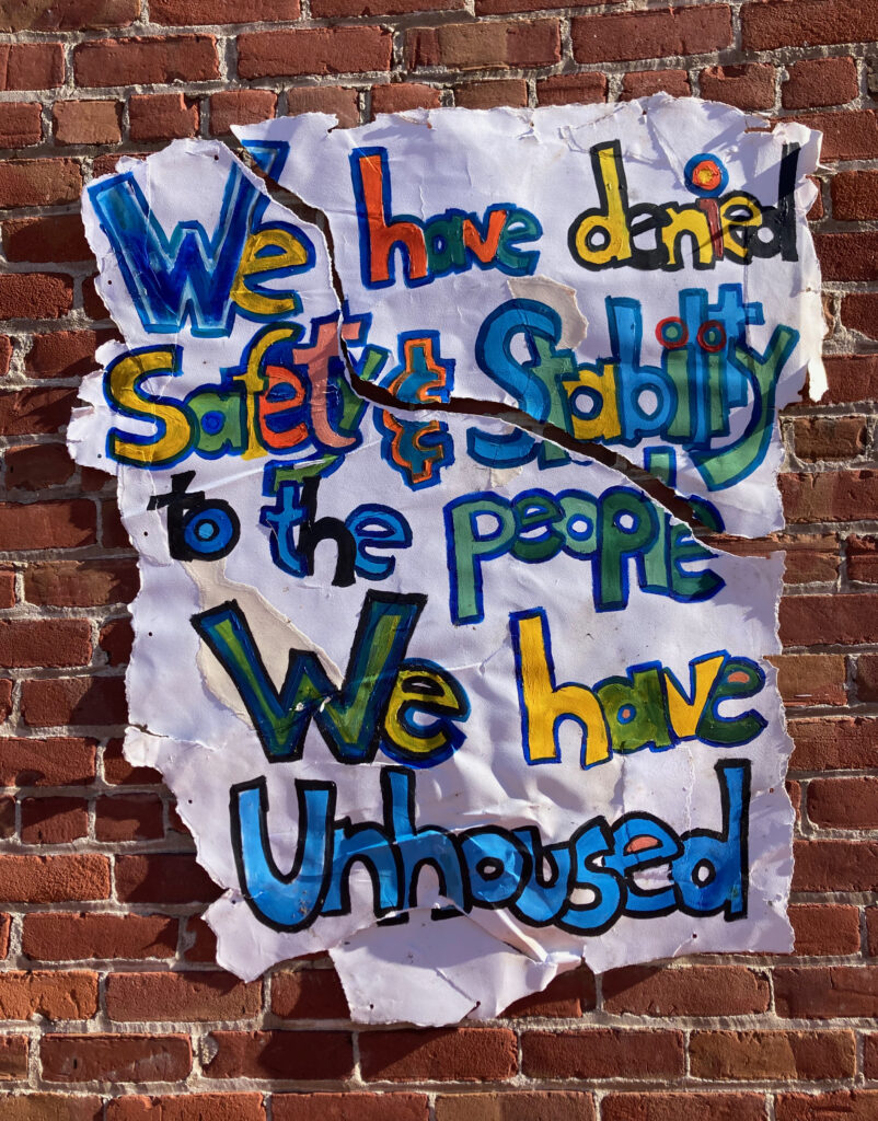 Torn poster showing damage from initial display. The poster reads "We have denied safety and stability to the people we have unhoused." The tear runs through the words "safety and stability."