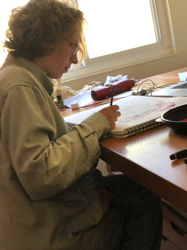 Shows female artist in profile painting at a table with a small paintbrush onto an art pad. Other art and painting supplies are nearby on the table.