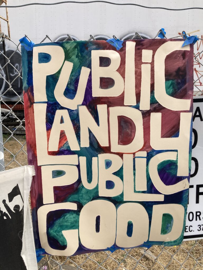 Hand printed poster hanging on fence reads "Public Land 4 Public Good" with dark multicolored background and cream letters from handcut stencils. Poster is hanging on chain link fence with blue painters tape. 