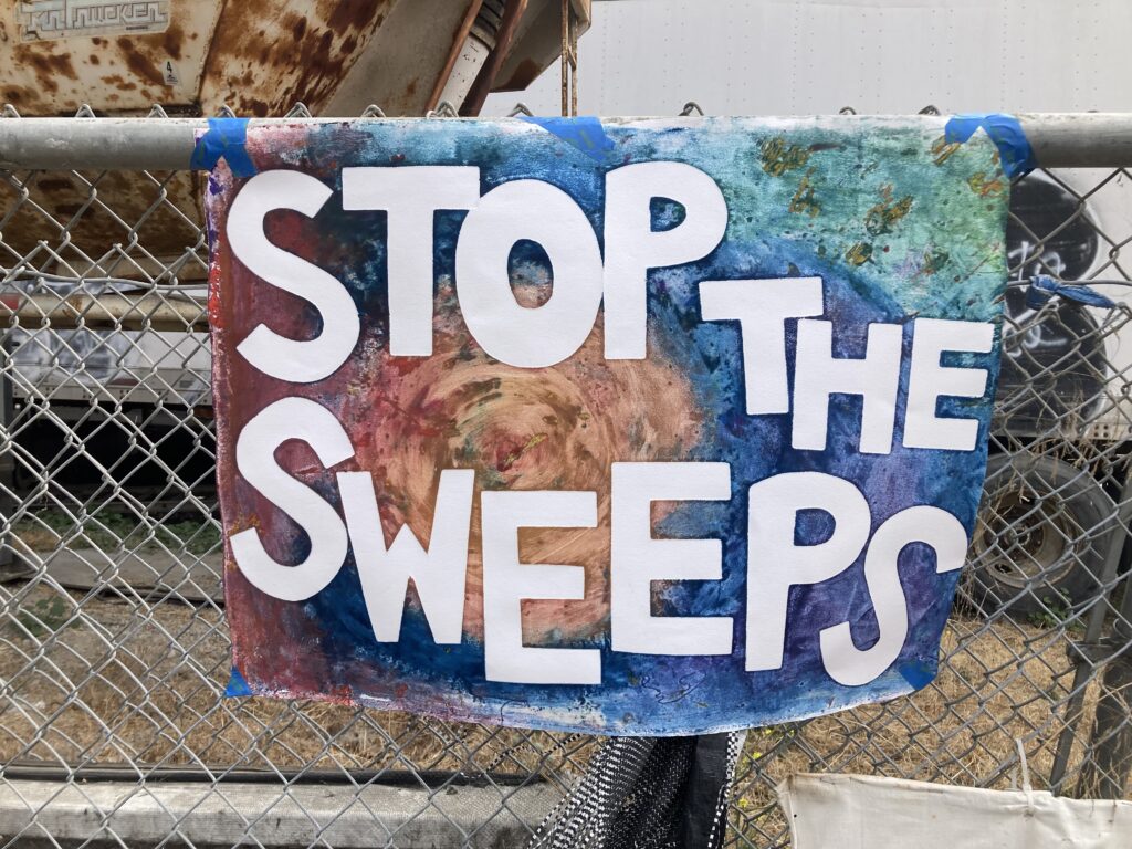 Poster with painterly background of blues, purples, greens and ochre with the text "Stop the Sweeps" in bold white contrast. Lettering is hand cut from stencils. Poster is hanging with blue tape on a chain link fence. 